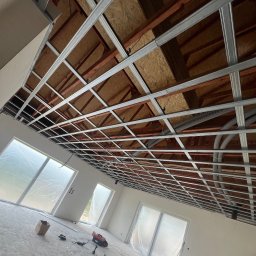 AllBud Usługi Ogólnobudowlane Łokietek Kamil - Widok na sufit z zamontowaną metalową konstrukcją pod płyty kartonowo-gipsowe, widoczne przewody wentylacyjne i oświetlenie, okna zabezpieczone folią malarską, narzędzia na podłodze.