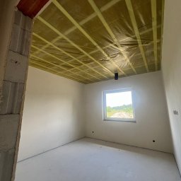 AllBud Usługi Ogólnobudowlane Łokietek Kamil - Wnętrze w stanie surowym: sufit z izolacją z folii i kraty z taśm, ściany w jasnym szarym kolorze, w tle okno z widokiem na zieleń.