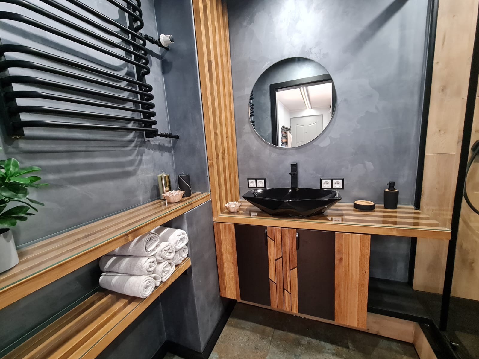 Nowoczesna łazienka z szarą ścianą o fakturze betonu, czarną umywalką w kształcie diamentu, okrągłym lustrem, drewnianymi elementami wykończeniowymi i czarnym grzejnikiem drabinkowym. Na półce leżą...