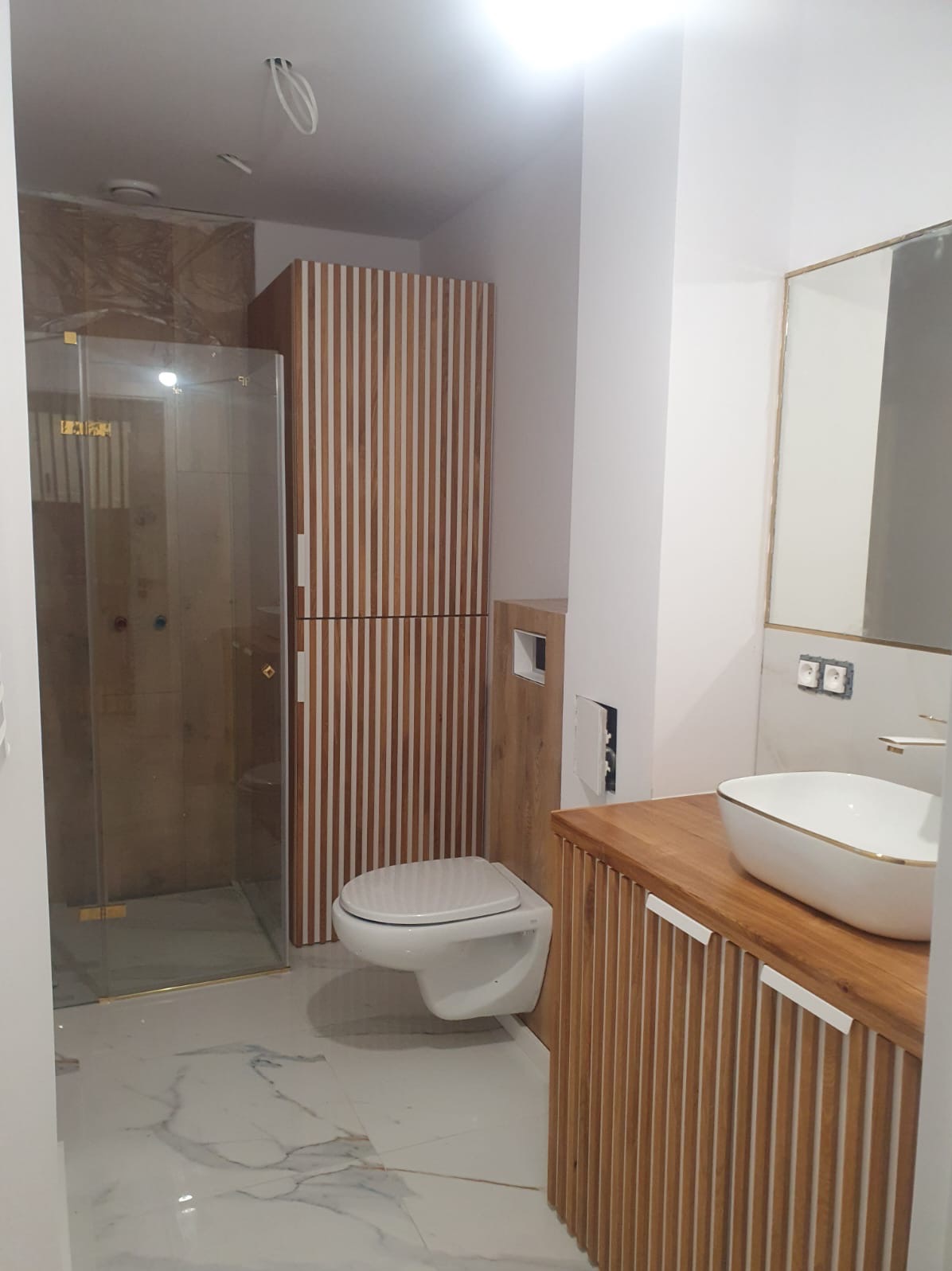 Nowoczesna łazienka z kabiną prysznicową, wiszącą toaletą i umywalką nablatową z drewnianą szafką w kolorze drewna z białymi akcentami.