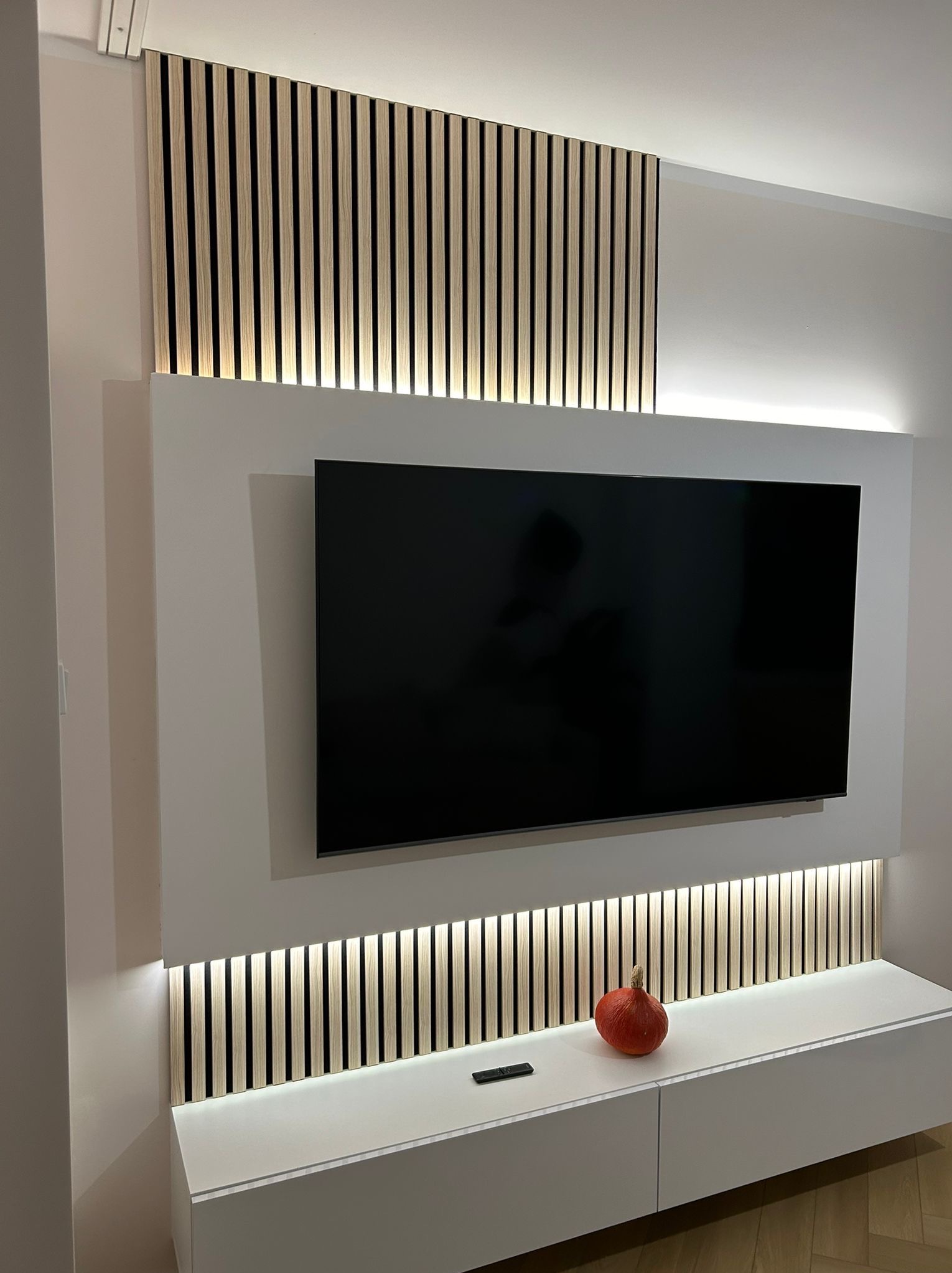 Ściana telewizyjna z podświetlanymi lamelami drewnianymi, biała szafka RTV z pomarańczową dekoracją w kształcie dyni.
