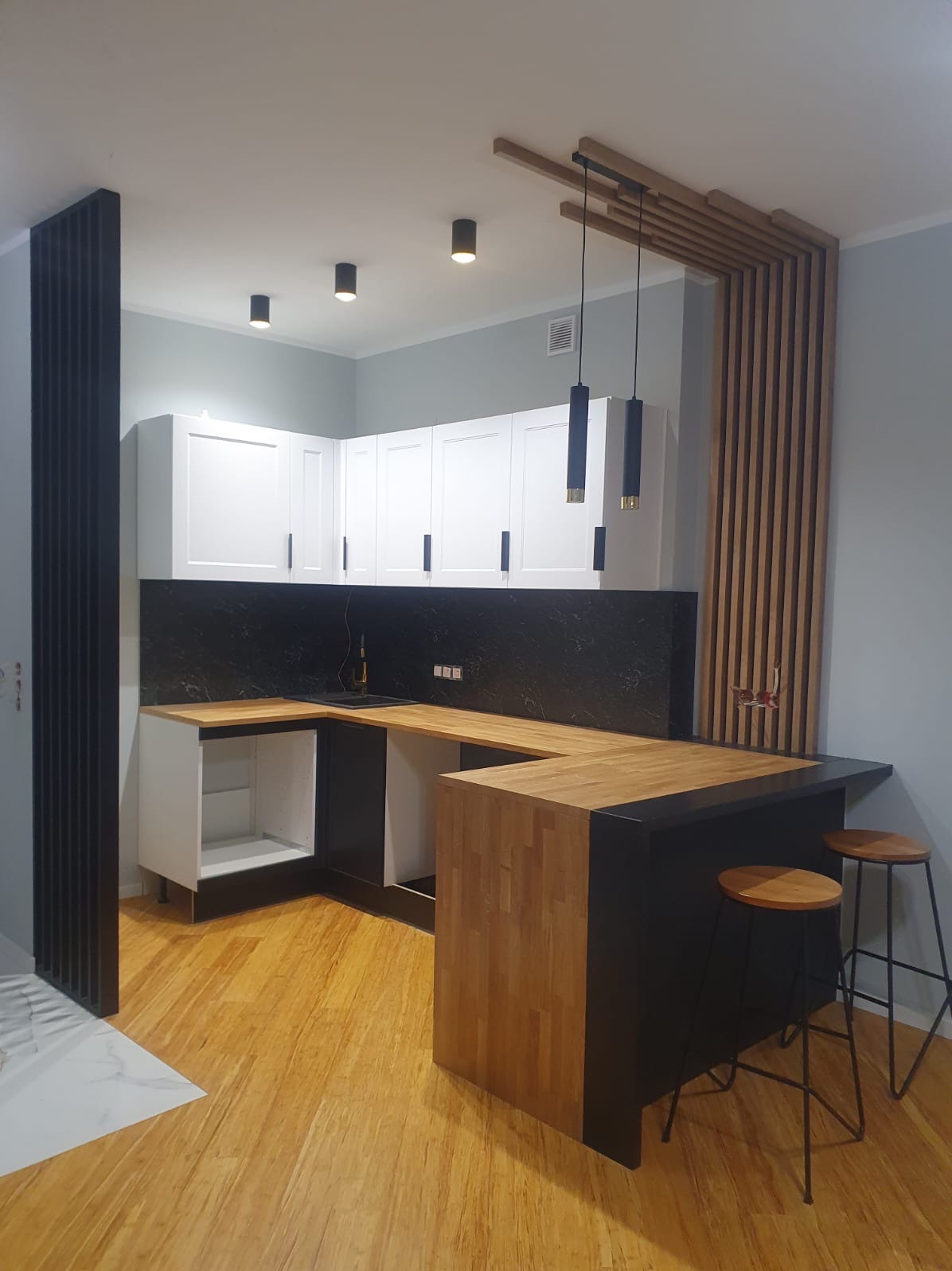 Nowoczesna kuchnia z drewnianym blatem, czarnymi elementami i białymi szafkami, uzupełniona drewnianymi lamelami na ścianie i minimalistycznymi lampami wiszącymi.