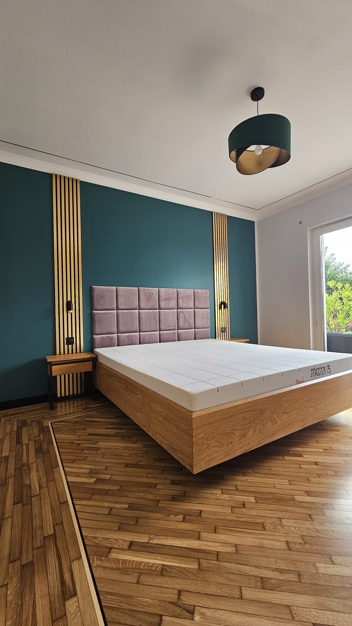 Sypialnia z łóżkiem na niskiej drewnianej ramie, wezgłowie z paneli w kolorze pudrowego różu, ściana w kolorze butelkowej zieleni z pionowymi złotymi panelami, parkiet w jodełkę.