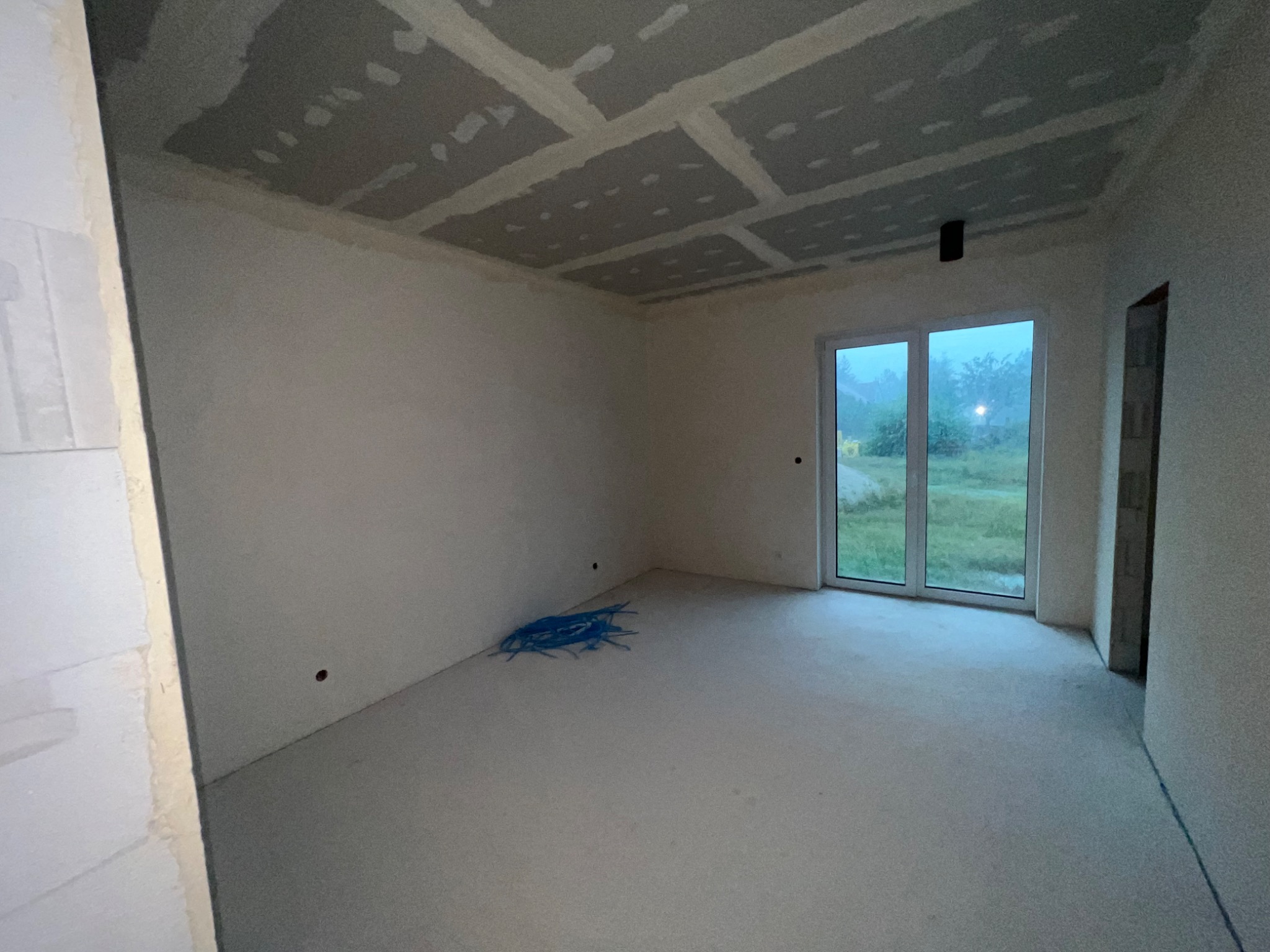 Pokój w stanie deweloperskim: surowe ściany, podłoga z wylewki, sufit z płyt kartonowo-gipsowych ze spoinami, okno balkonowe z widokiem na zewnątrz, niebieska wiązka kabli na podłodze.