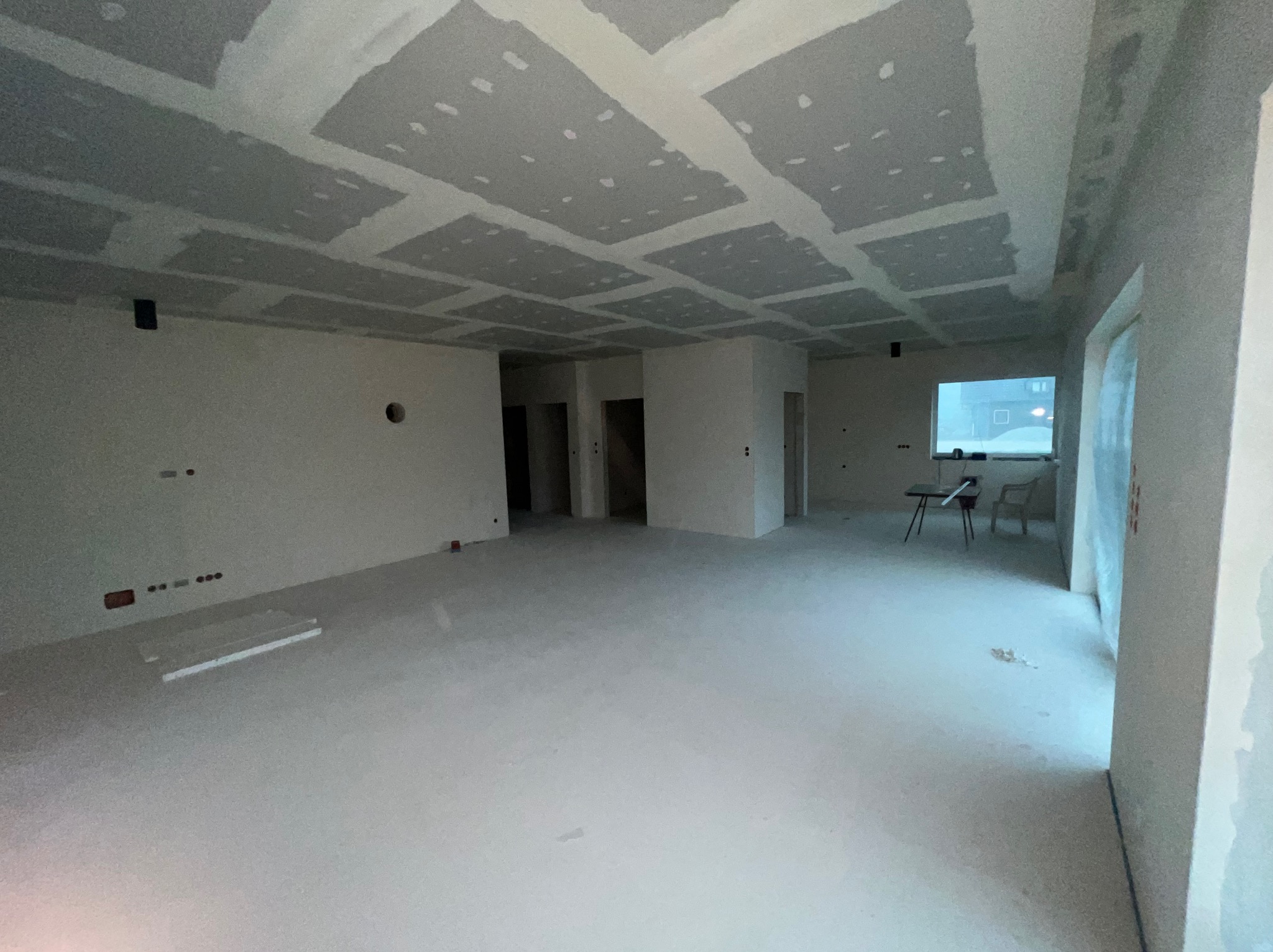 Przestronne wnętrze w trakcie prac wykończeniowych: sufit z płyt kartonowo-gipsowych z widocznymi łączeniami, ściany przygotowane pod malowanie, surowa podłoga, widoczne instalacje elektryczne...