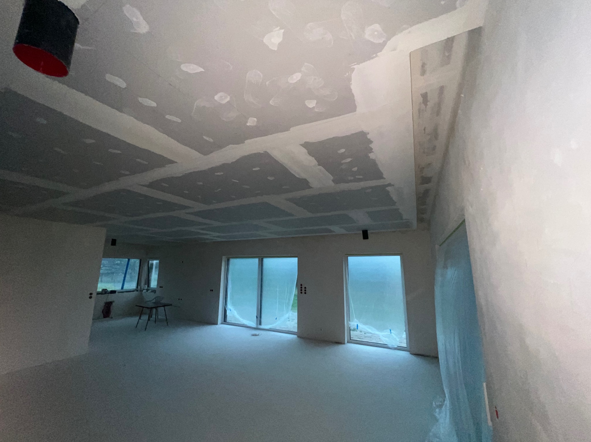 Wnętrze w trakcie prac wykończeniowych: sufit z płyt kartonowo-gipsowych z widocznymi łączeniami i punktowymi śladami szpachlowania, zabezpieczone folią okna, surowa podłoga.