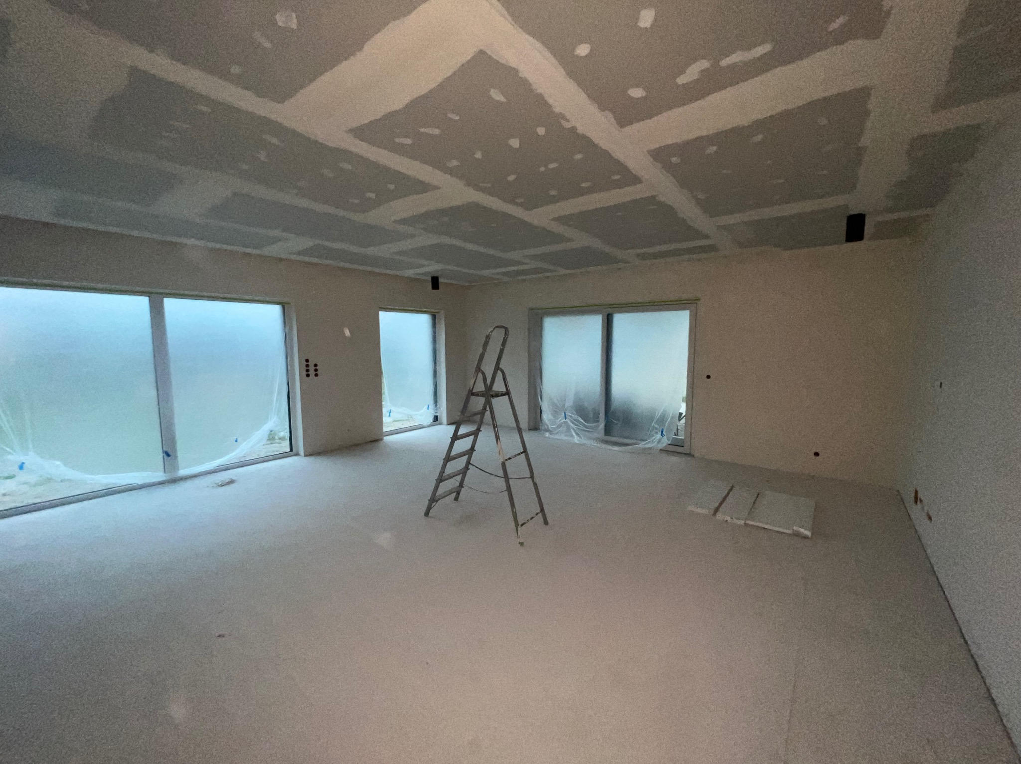 Pokój w trakcie prac wykończeniowych: ściany i sufit po szpachlowaniu, widoczne łączenia płyt karton-gips na suficie, drabina aluminiowa na środku, okna zabezpieczone folią malarską.