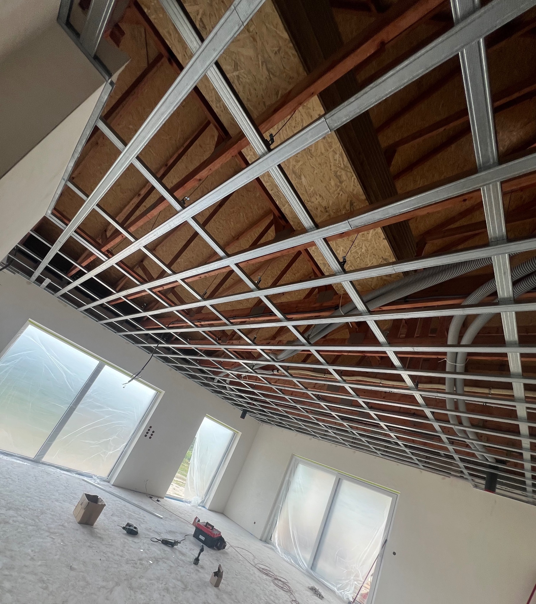 Widok na sufit z zamontowaną metalową konstrukcją pod płyty kartonowo-gipsowe, widoczne przewody wentylacyjne i oświetlenie, okna zabezpieczone folią malarską, narzędzia na podłodze.
