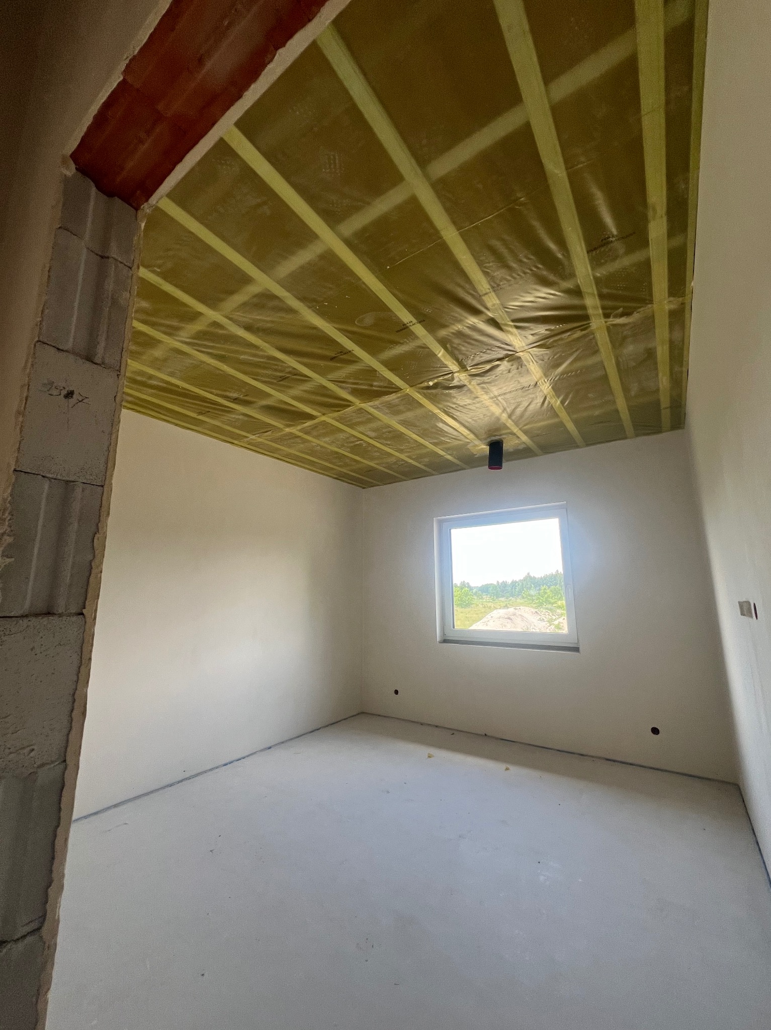 Wnętrze w stanie surowym: sufit z izolacją z folii i kraty z taśm, ściany w jasnym szarym kolorze, w tle okno z widokiem na zieleń.
