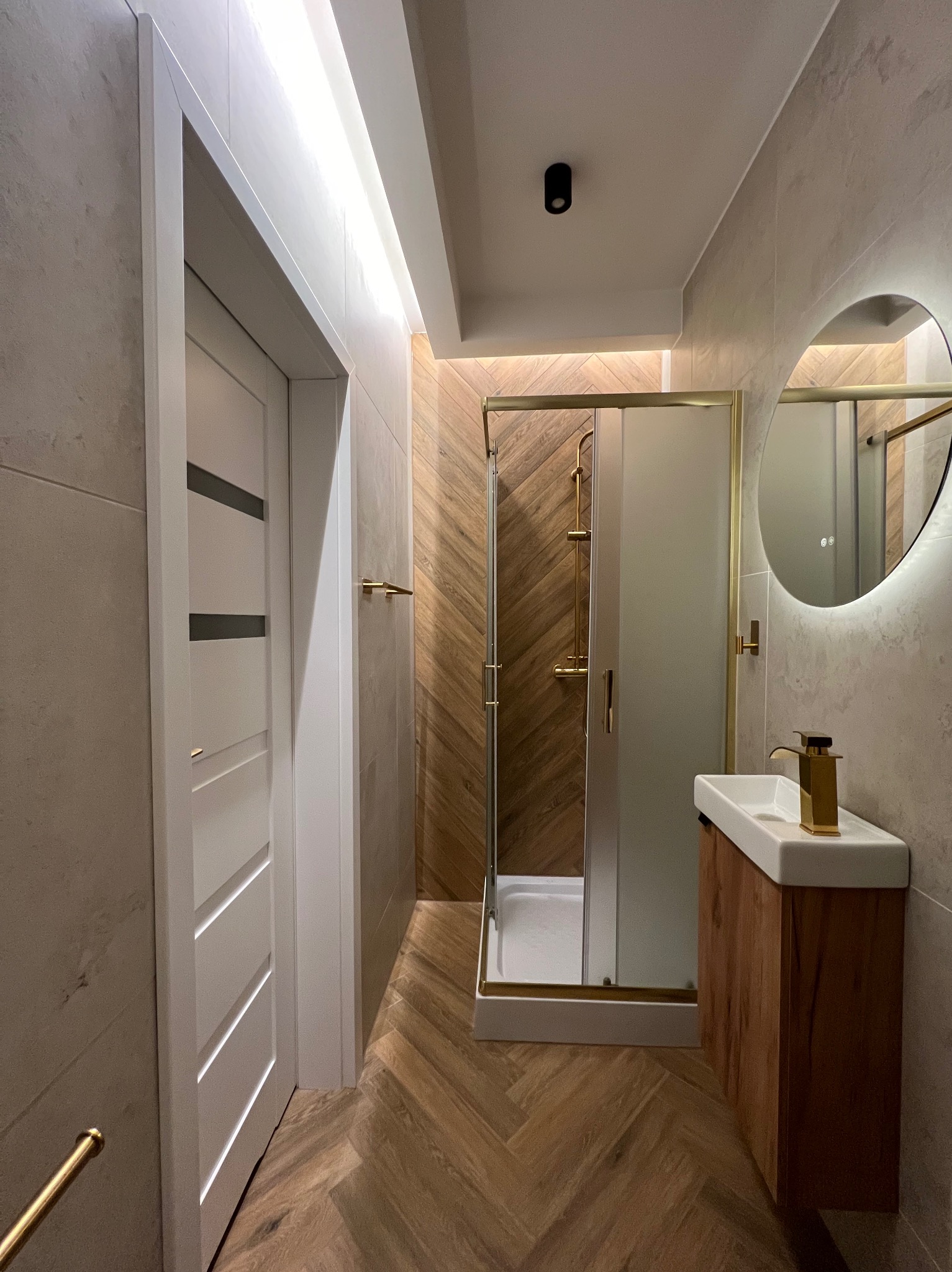 Nowoczesna łazienka z kabiną prysznicową w złotej ramie, umywalką na drewnianej szafce i okrągłym lustrem z podświetleniem LED. Parkiet ułożony w jodełkę.