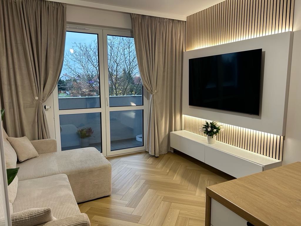 Nowoczesny salon z drewnianą ścianą, telewizorem i beżową sofą. Parkiet ułożony w jodełkę. Duże okno balkonowe z zasłonami w kolorze beżowym. Oświetlenie LED.