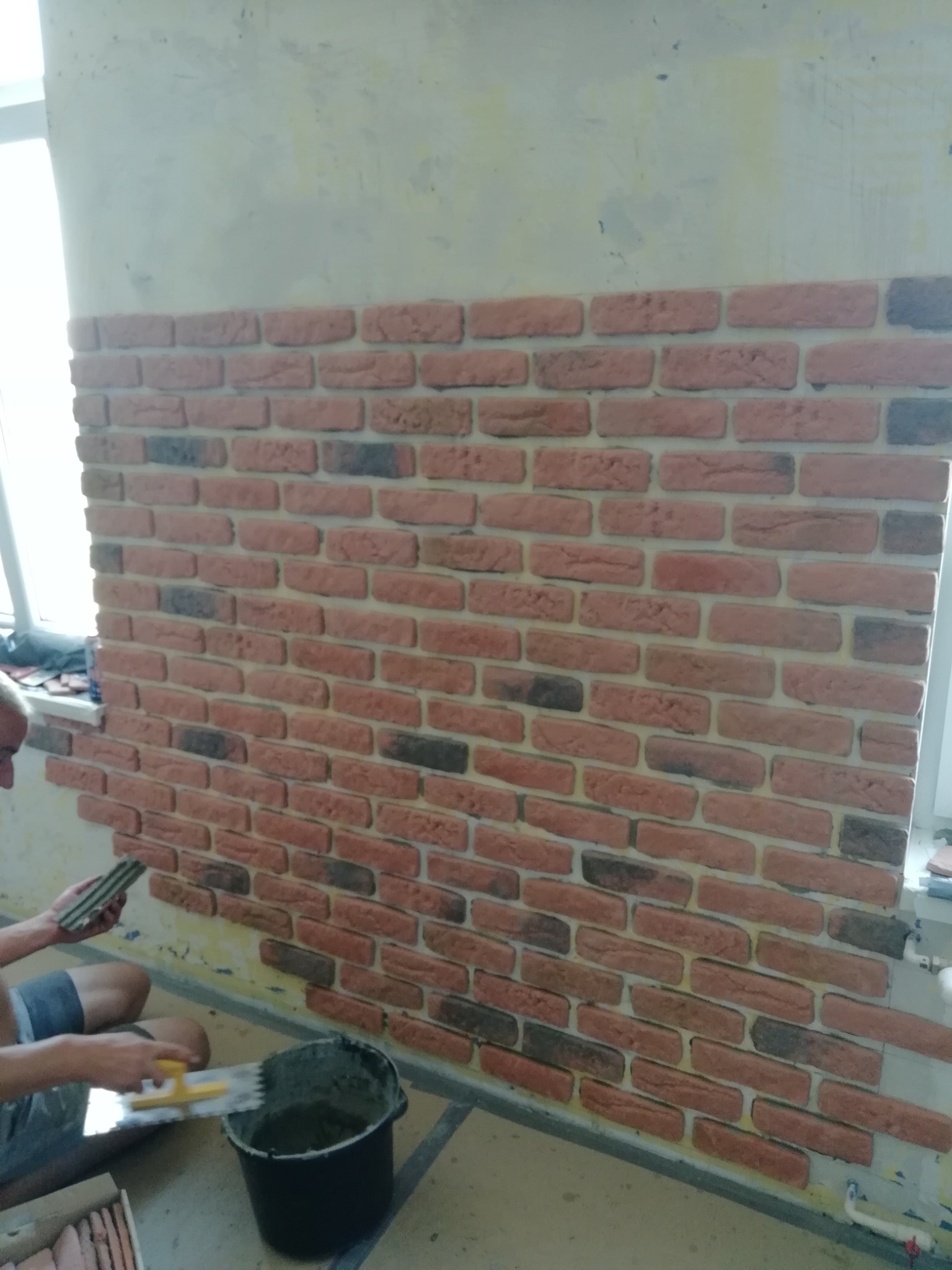 Fragment ściany z cegieł dekoracyjnych, w trakcie montażu. Widoczny pracownik z kielnią i wiadrem zaprawy, nakładający klej na płytkę. Częściowo wykończona powierzchnia z widocznymi różnicami...