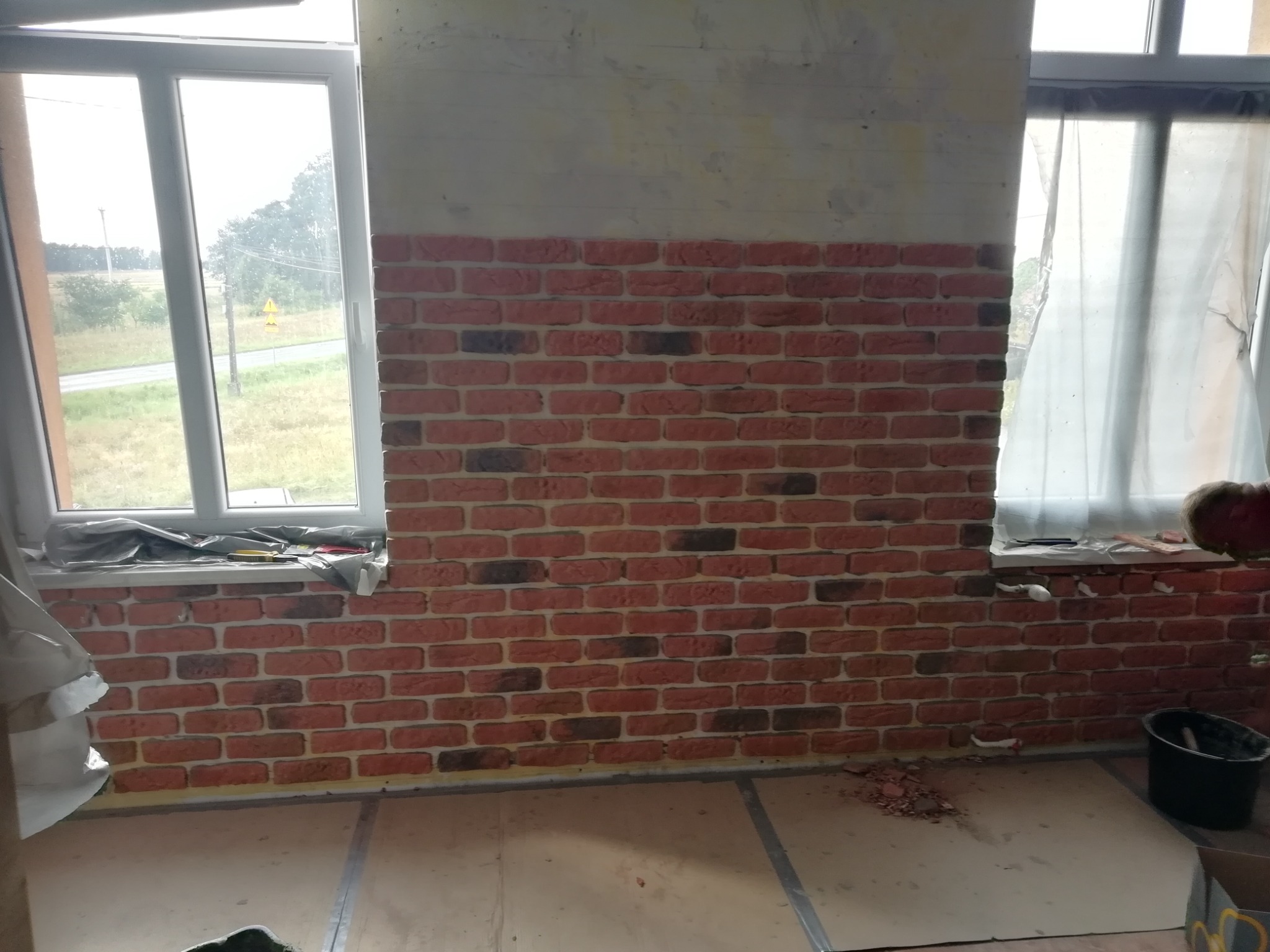 Ściana z cegieł dekoracyjnych w trakcie montażu, widoczne okna po bokach, pozostałości zaprawy na podłodze, surowy stan ściany powyżej cegieł.