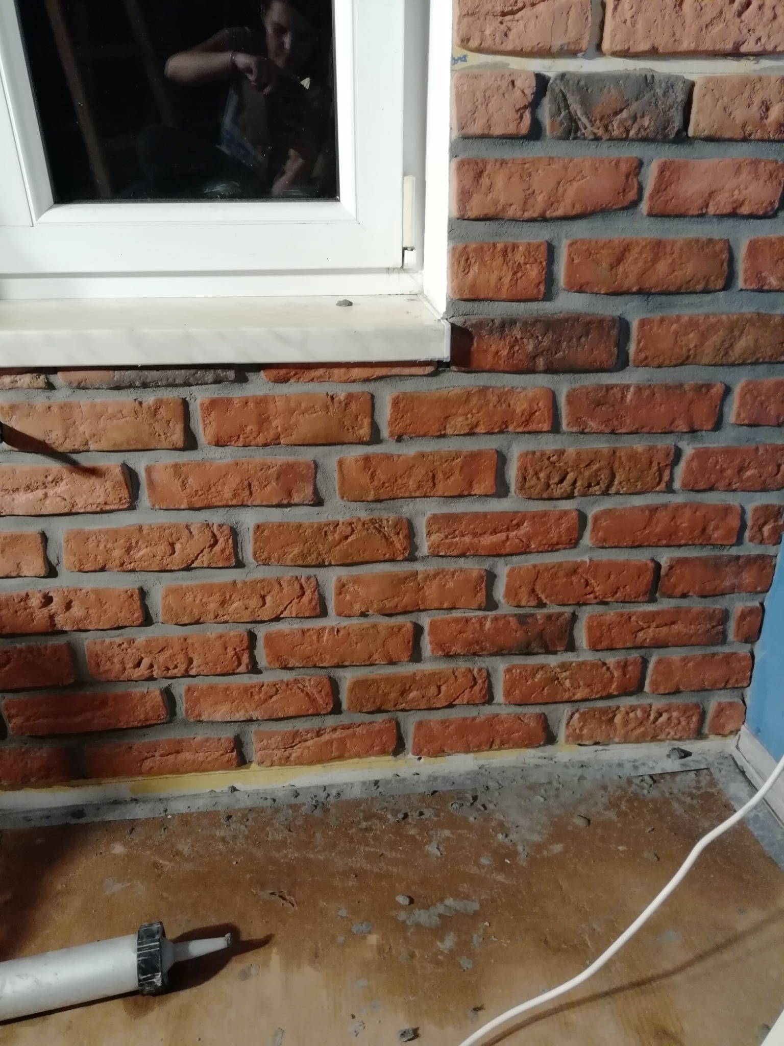 Fragment ściany z cegły klinkierowej pod oknem, widoczna fuga, narzędzie do fugowania na podłodze, odbicie osoby w szybie okna.