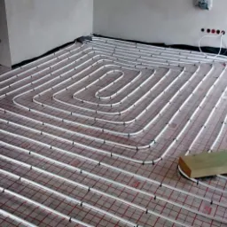 Montaż podłogowego ogrzewania wodnego z rur PEX na siatce z czerwonymi kwadratami, widoczne fragmenty ścian i wystające przewody elektryczne.