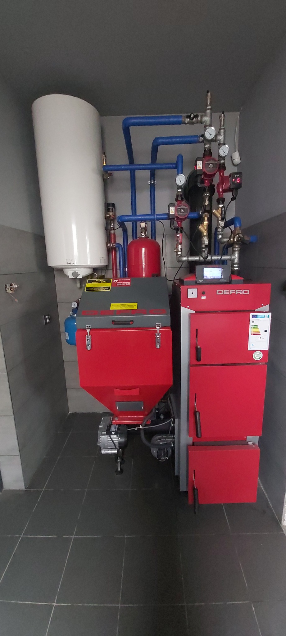 Czerwony piec z podajnikiem na ekogroszek marki Defro, z białym zasobnikiem na wodę, niebieskimi rurami instalacji CO, zamontowany w kotłowni z szarymi ścianami i ciemną podłogą z płytek.