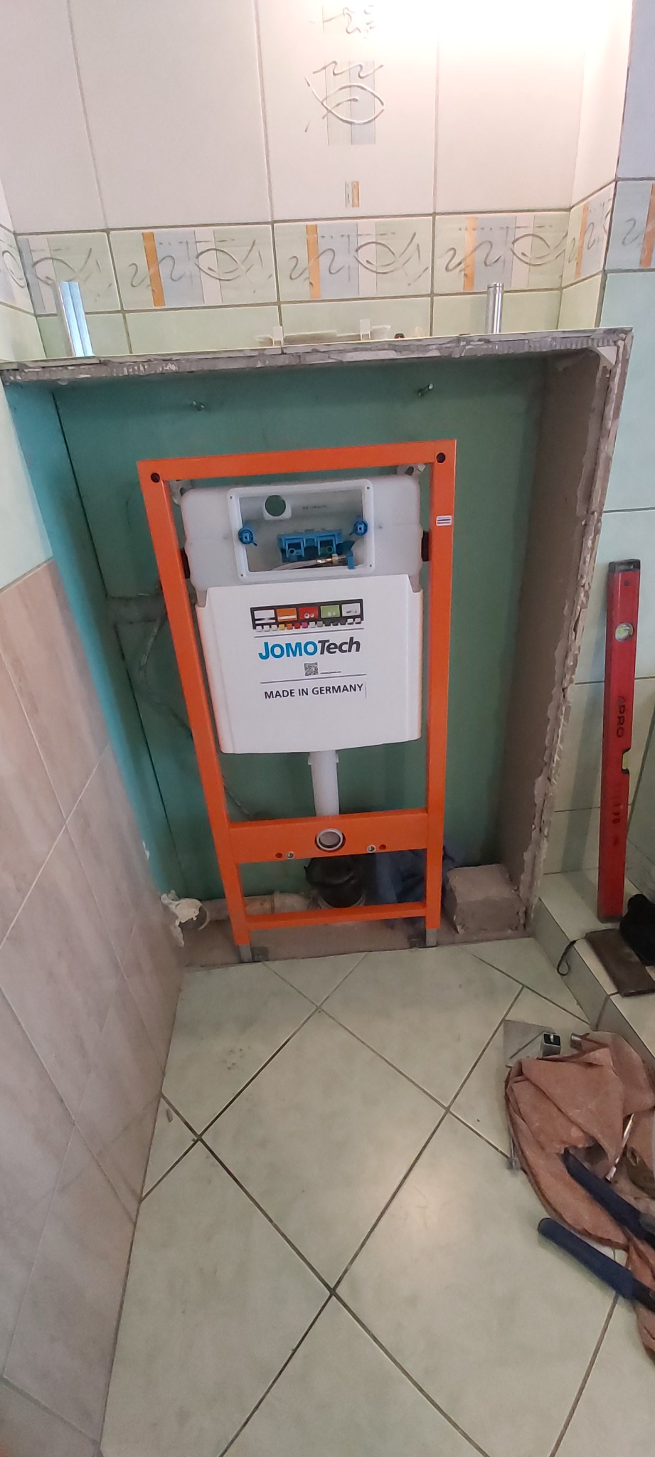 Stelaż podtynkowy JomoTech z widocznymi elementami instalacji wodno-kanalizacyjnej w trakcie montażu w obudowie z płyt gipsowo-kartonowych, poziomica oparta o ścianę.