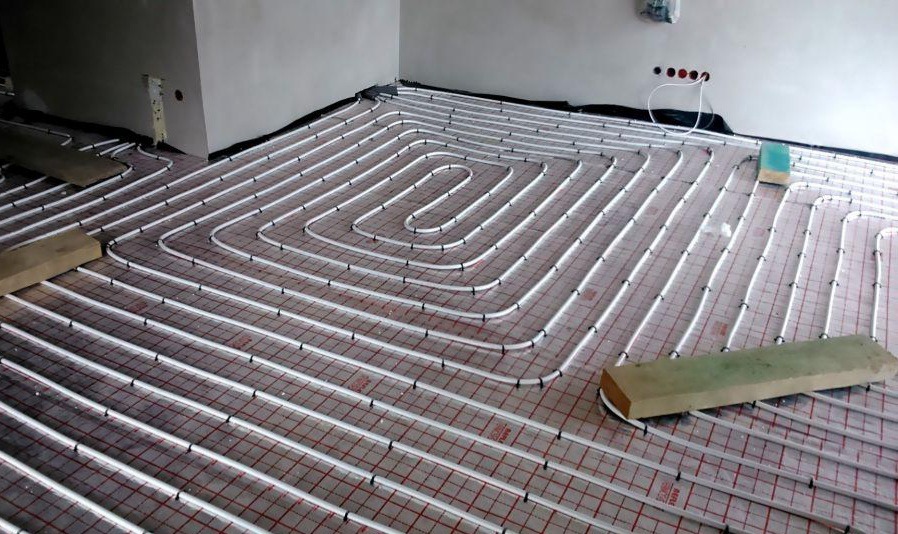 Montaż podłogowego ogrzewania wodnego z rur PEX na siatce z czerwonymi kwadratami, widoczne fragmenty ścian i wystające przewody elektryczne.