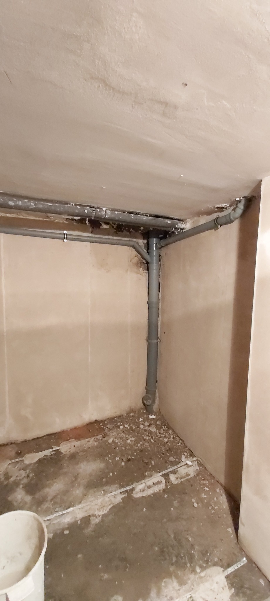 Szare rury kanalizacyjne pod sufitem w rogu pomieszczenia w trakcie remontu, widoczne ślady po pracach na ścianach i podłodze.