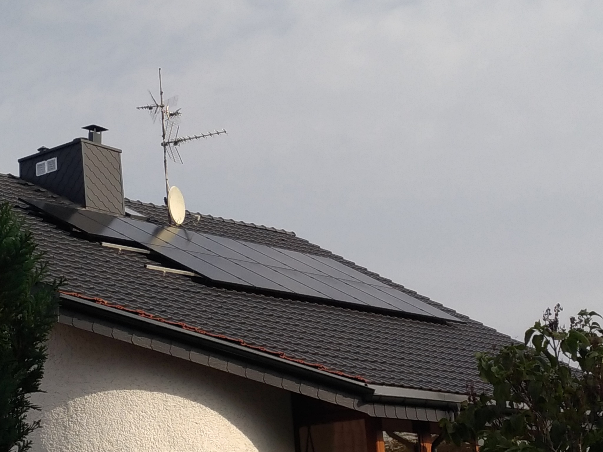 Panele słoneczne na dachu domu jednorodzinnego z anteną satelitarną i kominem w tle.