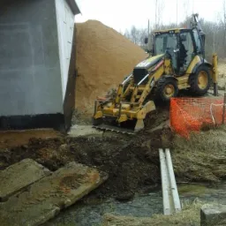 Żółta koparko-ładowarka Caterpillar 432E wykonuje prace ziemne przy betonowym filarze mostu, z widoczną stertą piasku w tle i małym ciekiem wodnym w pierwszym planie.