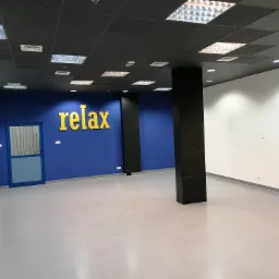Wnętrze biura po remoncie: Świeżo pomalowana niebieska ściana z napisem 'relax' w kolorze żółtym, czarny filar, szara podłoga z płytek, białe ściany i sufit z panelami rastrowymi.