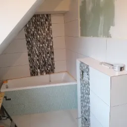 Łazienka w trakcie remontu na poddaszu, widoczna wanna obłożona mozaiką, ściany z białych płytek, fragment ściany z przygotowaną powierzchnią pod malowanie oraz zabudowa podtynkowego systemu WC...