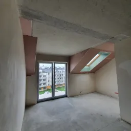 Wnętrze poddasza z oknem dachowym w trakcie zabudowy płytami gipsowo-kartonowymi; widoczne surowe ściany i betonowa podłoga, na zewnątrz widok na osiedle mieszkaniowe.