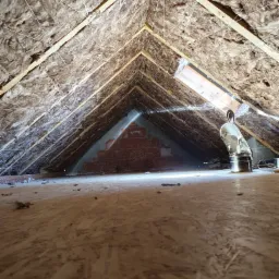 Poddasze z ociepleniem z wełny mineralnej, widoczne drewniane krokwie, metalowy przewód wentylacyjny i fragment muru z cegieł w głębi.