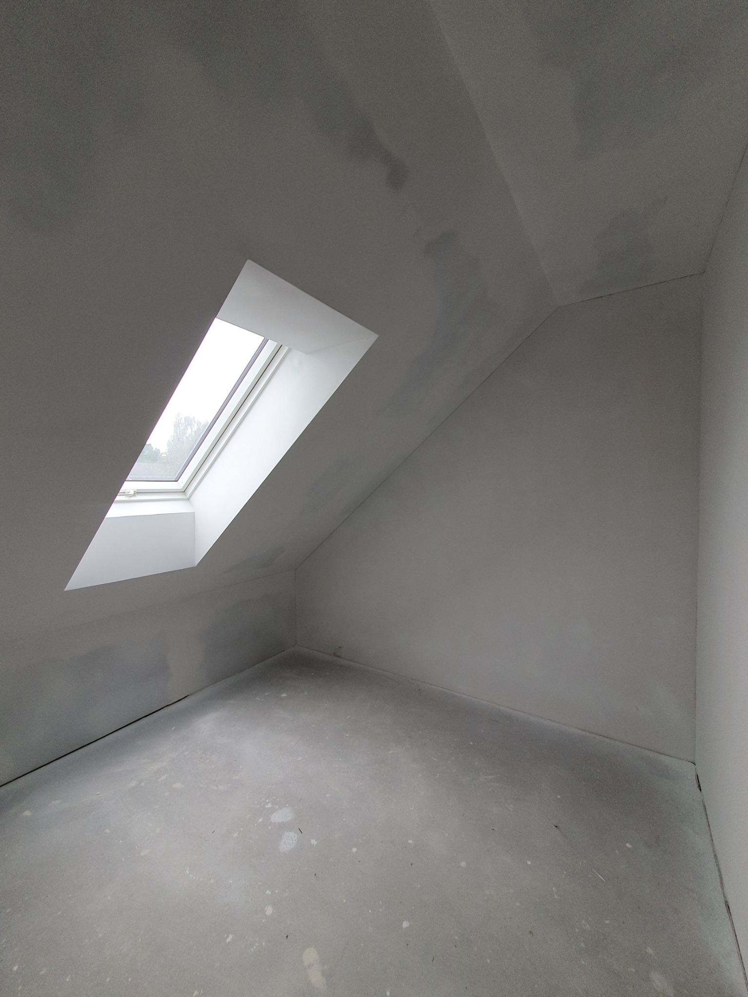 Poddasze ze skośnym sufitem i oknem dachowym w trakcie prac wykończeniowych, widoczne ślady szpachlowania i przygotowania do malowania, surowy beton na podłodze.