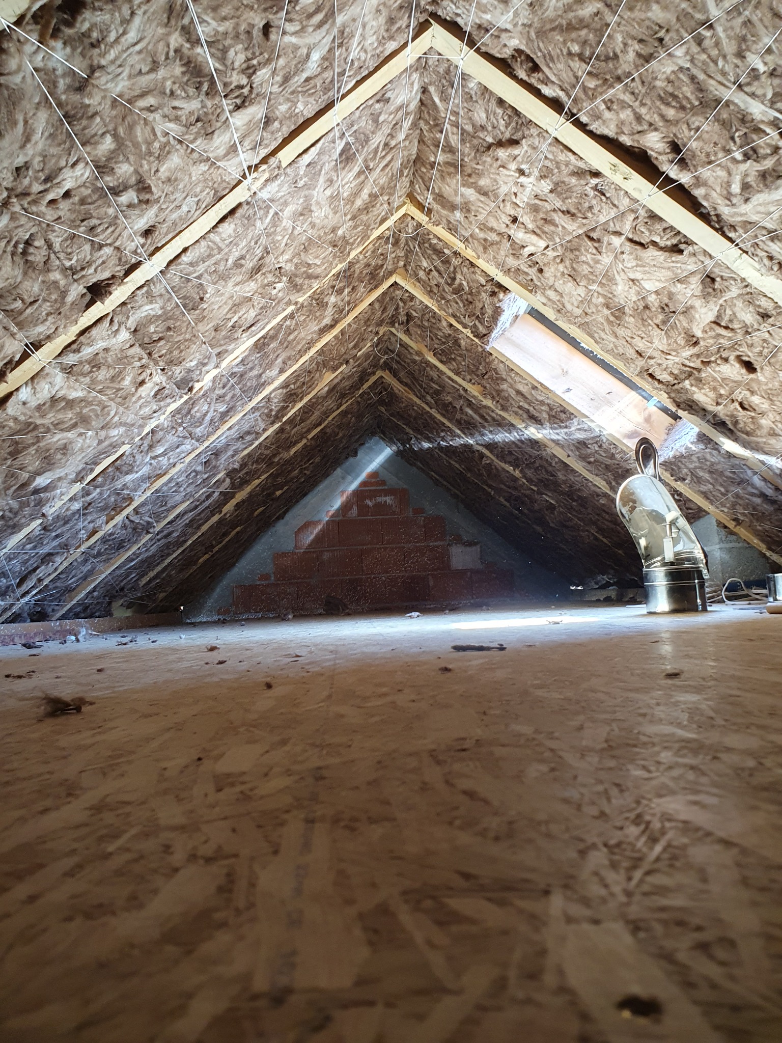 Poddasze z ociepleniem z wełny mineralnej, widoczne drewniane krokwie, metalowy przewód wentylacyjny i fragment muru z cegieł w głębi.