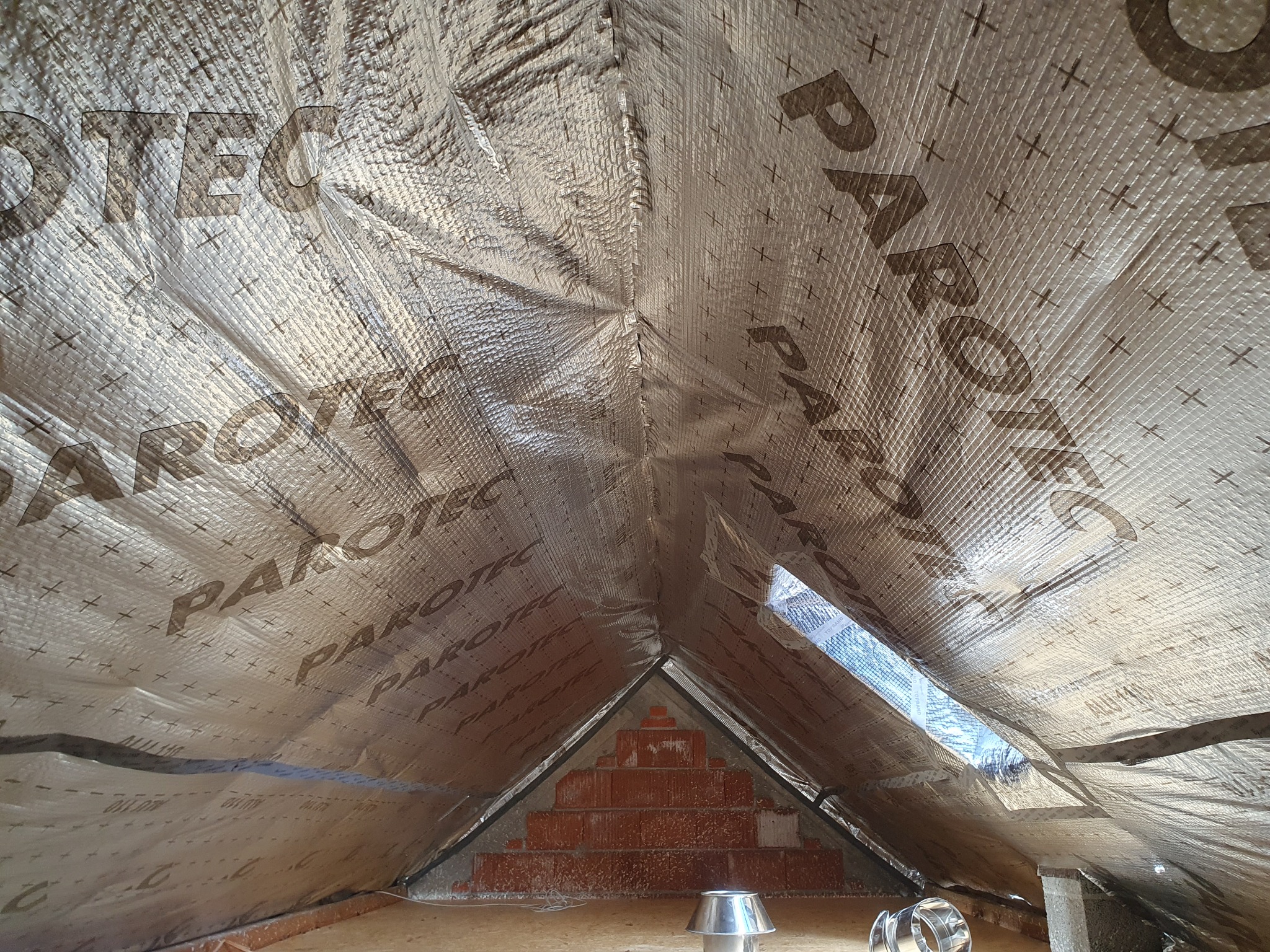 Poddasze z widoczną izolacją termiczną marki Parotec, folia aluminiowa z nadrukiem, ceglany komin w szczycie dachu, wentylacja kominkowa na podłodze.