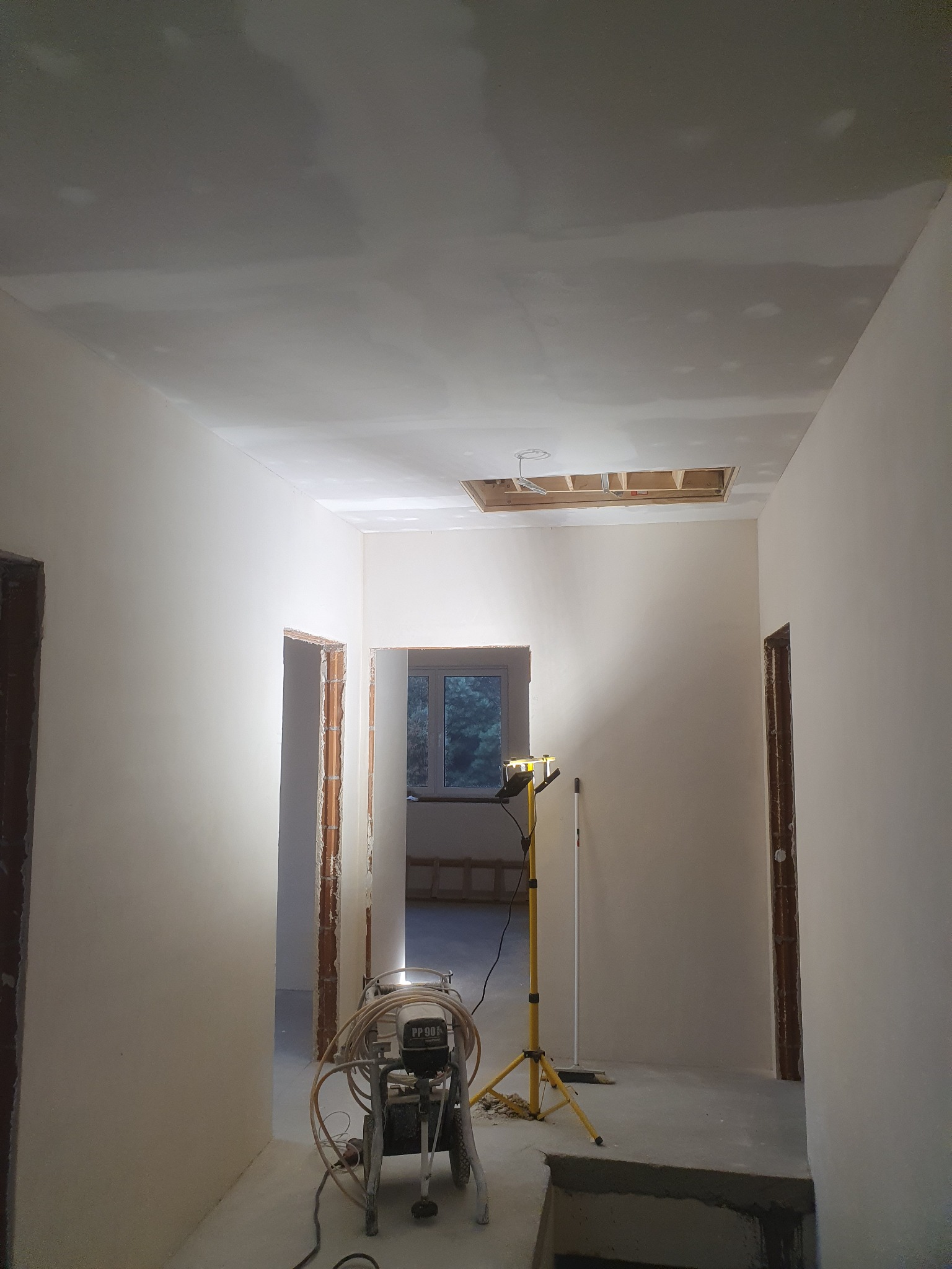 Wnętrze w trakcie prac wykończeniowych: świeżo wyszpachlowane ściany i sufit z widocznym otworem rewizyjnym, profesjonalny agregat malarski na podłodze i lampa budowlana oświetlająca pomieszczenie.