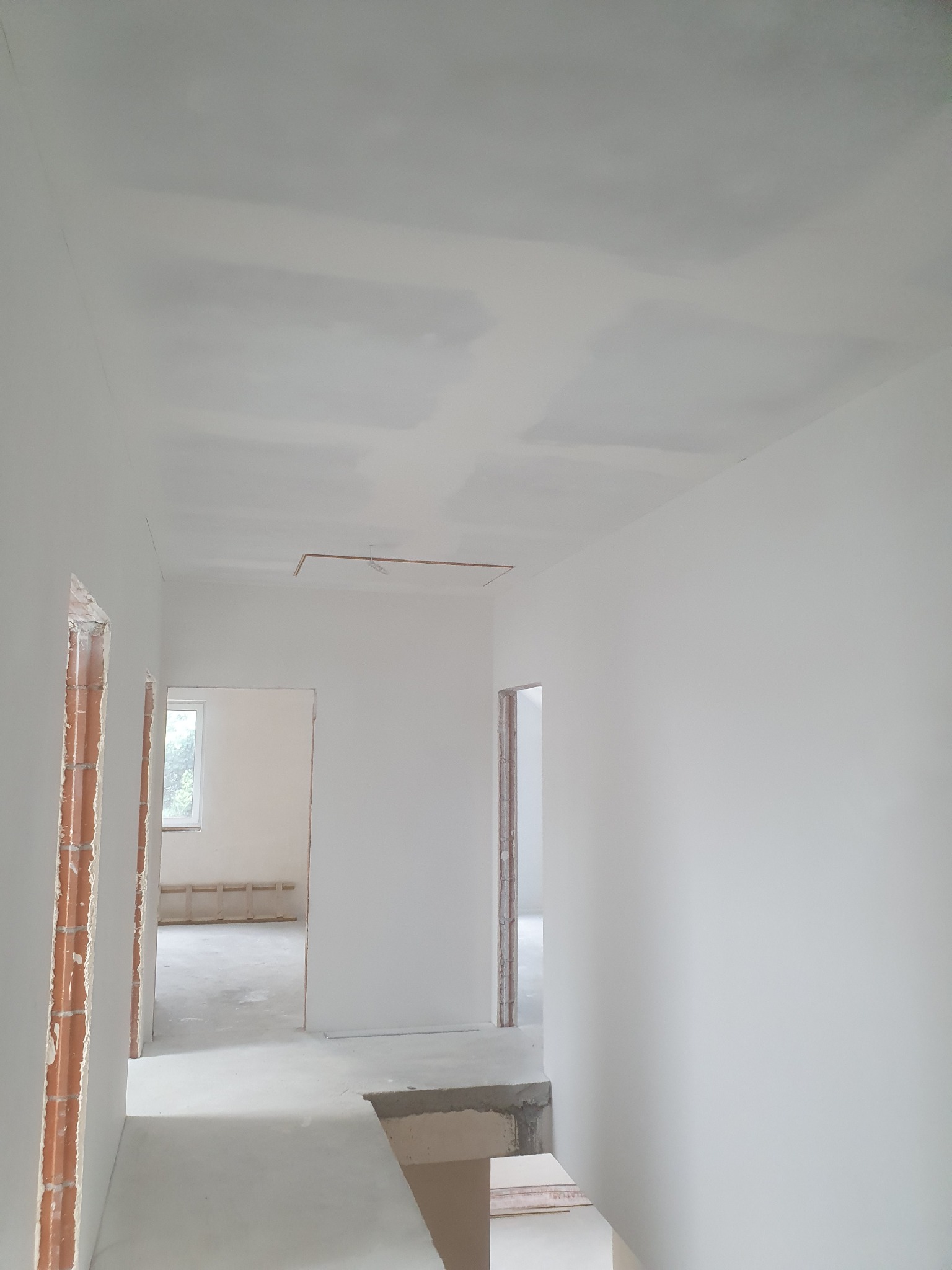 Wnętrze w trakcie wykańczania: surowe ściany i strop po szpachlowaniu, widoczne niedoskonałości i różnice w odcieniach bieli, fragment instalacji elektrycznej na suficie, ceglane ościeżnice drzwi.