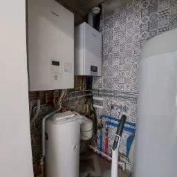Dwa kotły grzewcze, marki Panasonic i Vaillant, zamontowane w pomieszczeniu z ozdobnymi płytkami ściennymi, widoczne rury i instalacje.