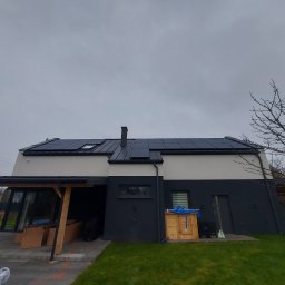 EcoPower+ Mateusz Kempiński - Dwukondygnacyjny dom z panelami na dachu, drewniana wiata garażowa w trakcie budowy, drewniana obudowa pompy ciepła z niebieskim wężem na wierzchu, pochmurne niebo.