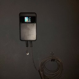 EcoPower+ Mateusz Kempiński - Czarna, naścienna stacja ładowania pojazdów elektrycznych z wyświetlaczem, kablem i wtyczką, zamontowana na ciemnej ścianie.