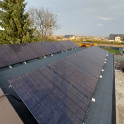 EcoPower+ Mateusz Kempiński - Rząd paneli słonecznych na płaskim dachu pokrytym papą, widok z góry na tle zabudowy jednorodzinnej i pochmurnego nieba.