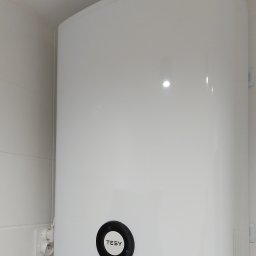 EcoPower+ Mateusz Kempiński - Biały, wiszący bojler elektryczny marki Tesy na tle białych kafelków, z widocznym przewodem zasilającym i podłączeniem hydraulicznym.