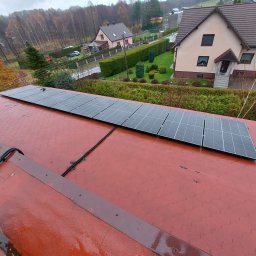 EcoPower+ Mateusz Kempiński - Rząd paneli słonecznych na czerwonym, mokrym dachu domu jednorodzinnego w pochmurny dzień. Widok z góry, na drugim planie zabudowa mieszkalna i drzewa.