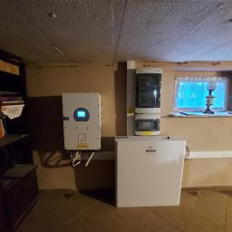 EcoPower+ Mateusz Kempiński - Biały inwerter z wyświetlaczem, skrzynka bezpiecznikowa i magazyn energii Encor w pomieszczeniu z oknem i lampą na parapecie.