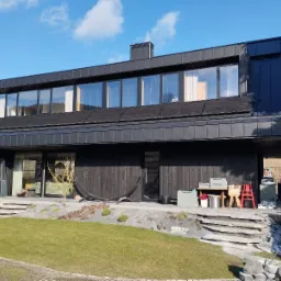 Nowoczesny dom z ciemnego drewna z panelami słonecznymi na dachu, widoczne z zewnątrz, z elementami wykończeniowymi i narzędziami na tarasie.