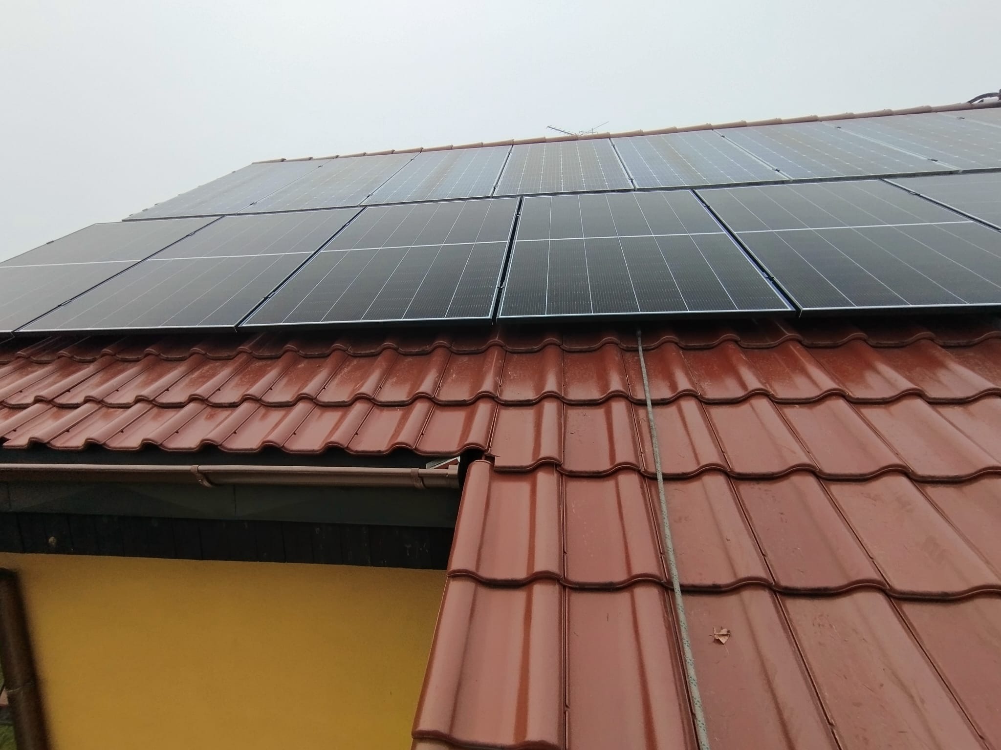 Czerwony dach z dachówki ceramicznej, na którym zamontowano ciemne panele słoneczne w pochmurny dzień. Widoczny maszt antenowy.