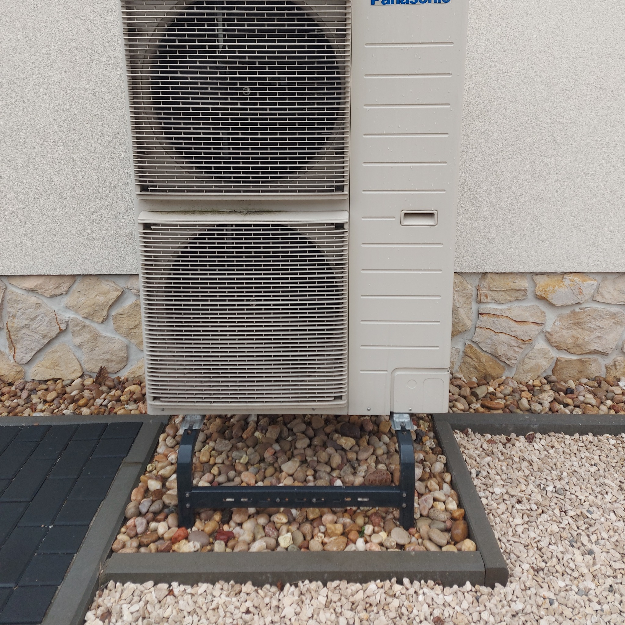 Zewnętrzna jednostka pompy ciepła Panasonic zamontowana na metalowym stelażu, otoczona ozdobnymi kamieniami w dwóch kolorach, przy ścianie budynku z kamienną podmurówką.