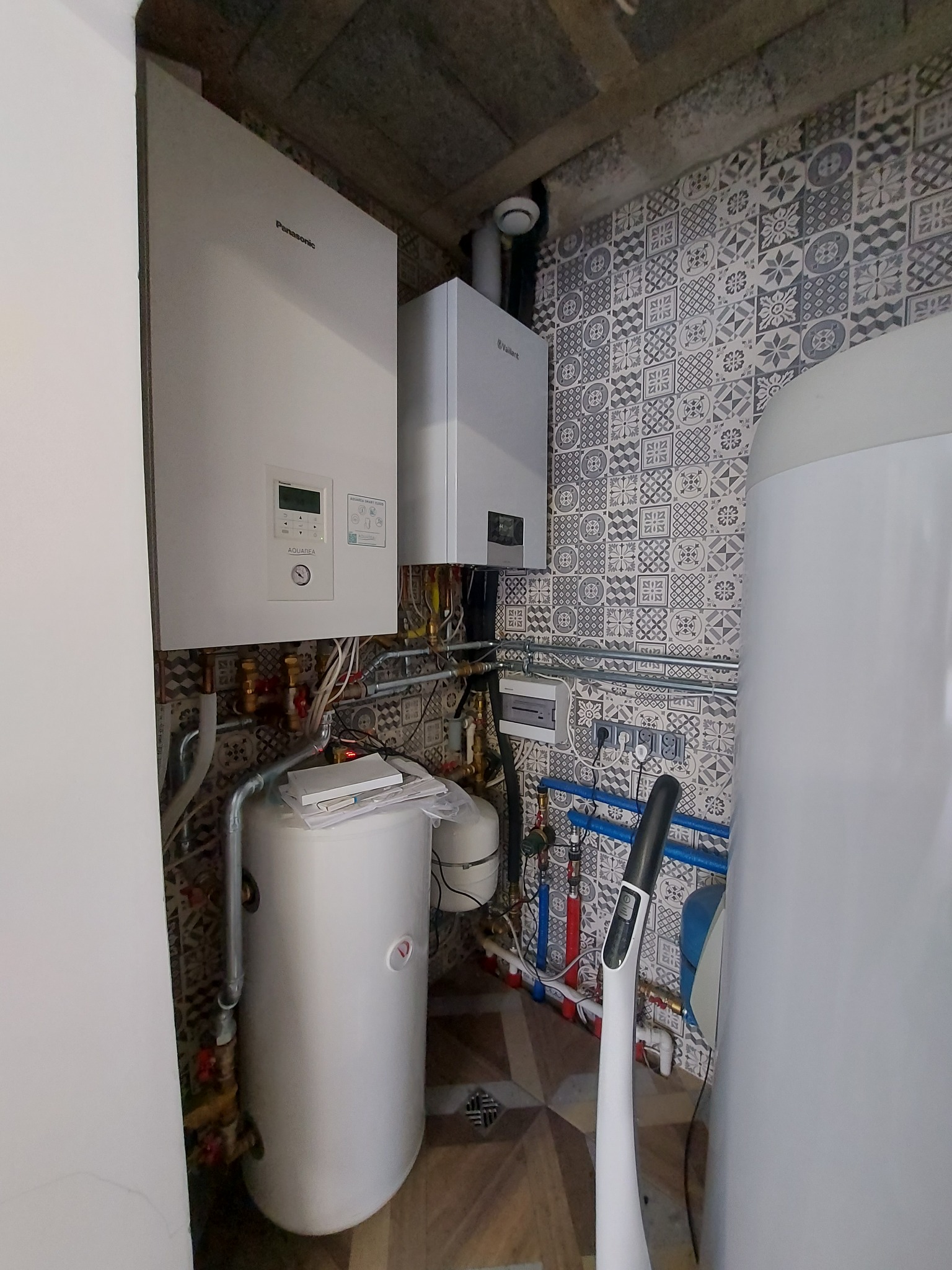 Dwa kotły grzewcze, marki Panasonic i Vaillant, zamontowane w pomieszczeniu z ozdobnymi płytkami ściennymi, widoczne rury i instalacje.