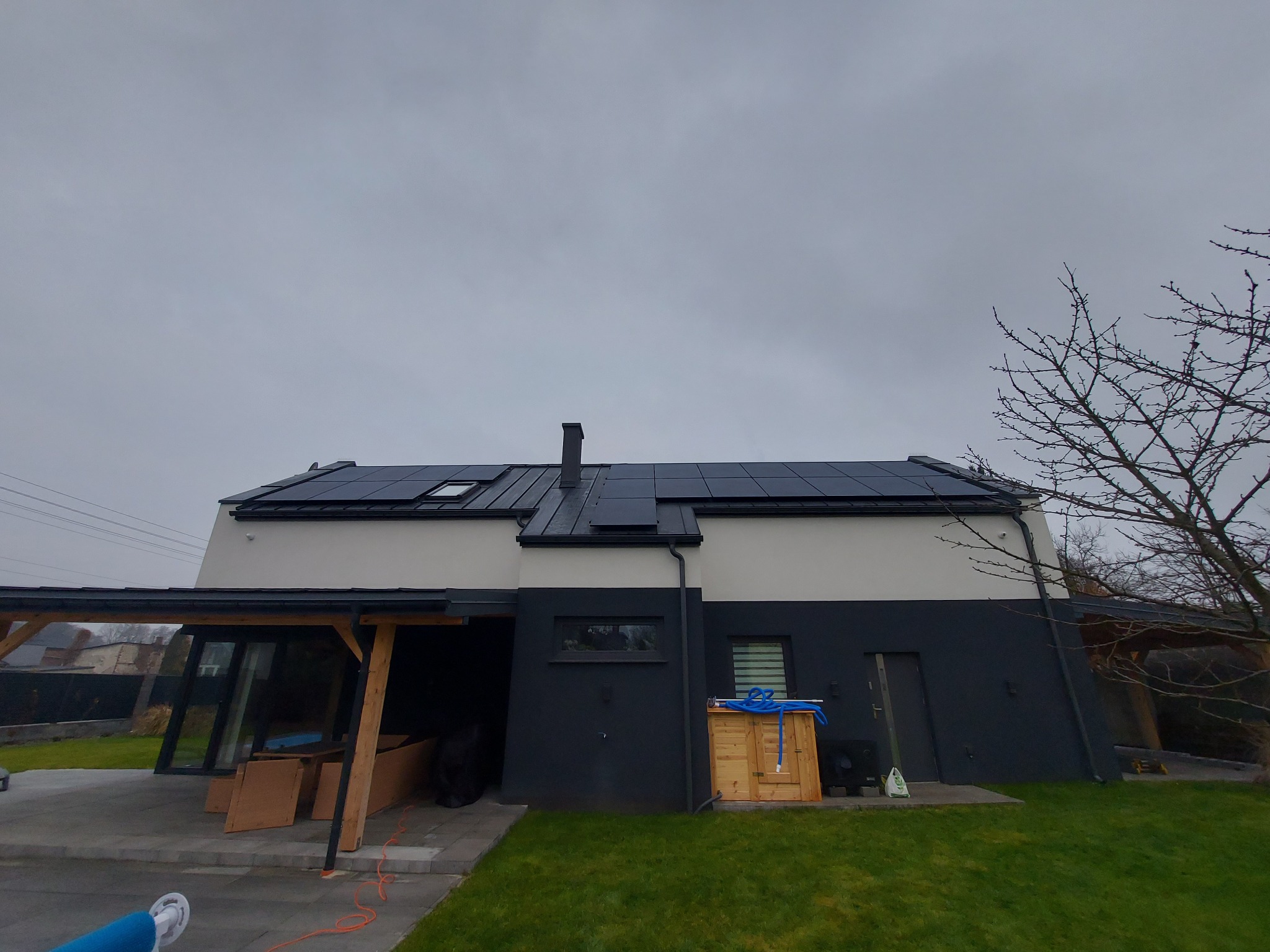 Dwukondygnacyjny dom z panelami na dachu, drewniana wiata garażowa w trakcie budowy, drewniana obudowa pompy ciepła z niebieskim wężem na wierzchu, pochmurne niebo.