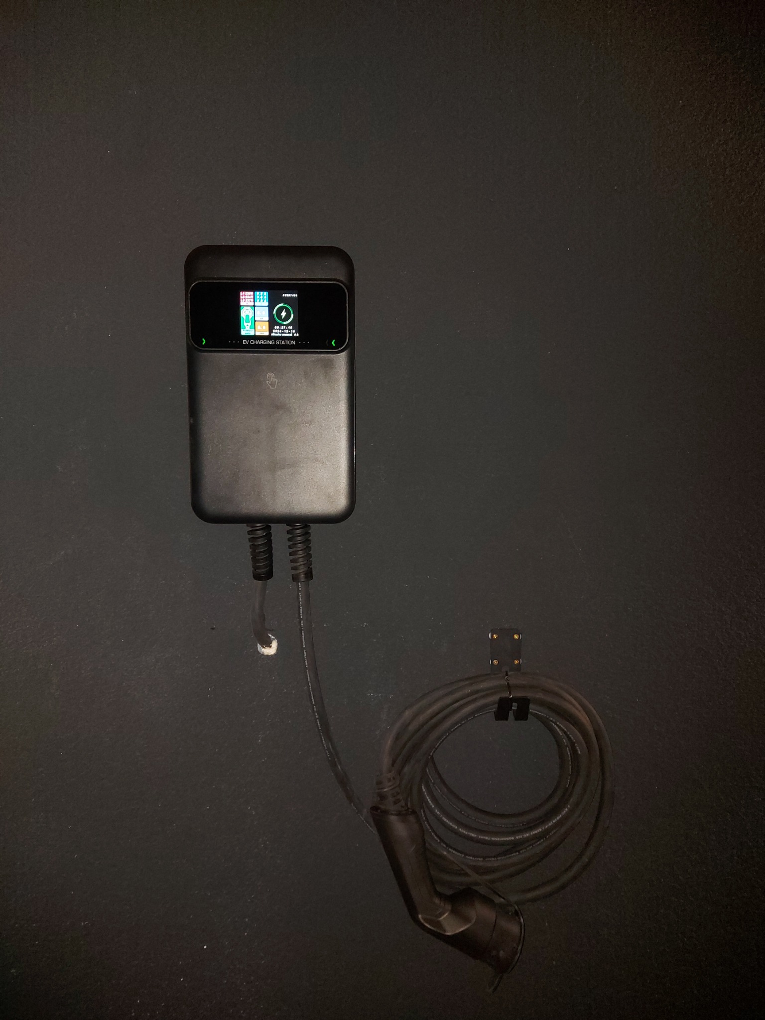 Czarna, naścienna stacja ładowania pojazdów elektrycznych z wyświetlaczem, kablem i wtyczką, zamontowana na ciemnej ścianie.