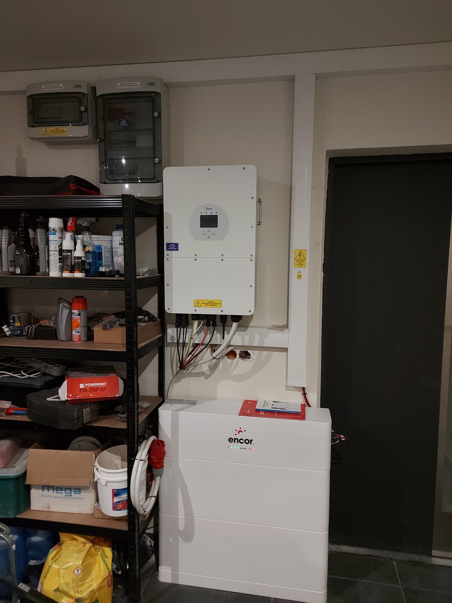 Biały inwerter Deye i magazyn energii Encor zamontowane na ścianie w pomieszczeniu gospodarczym, obok regału z różnymi przedmiotami i skrzynki elektryczne na górze.