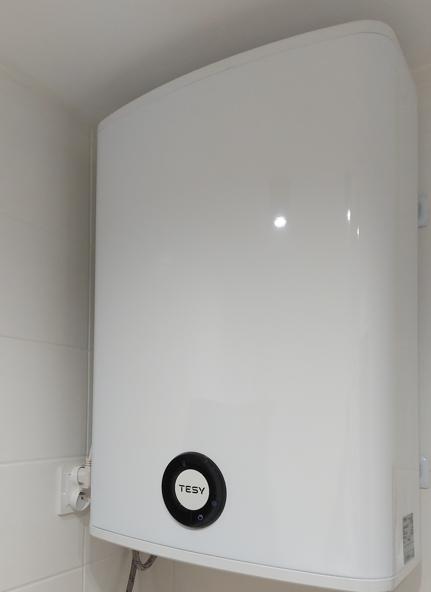 Biały, wiszący bojler elektryczny marki Tesy na tle białych kafelków, z widocznym przewodem zasilającym i podłączeniem hydraulicznym.
