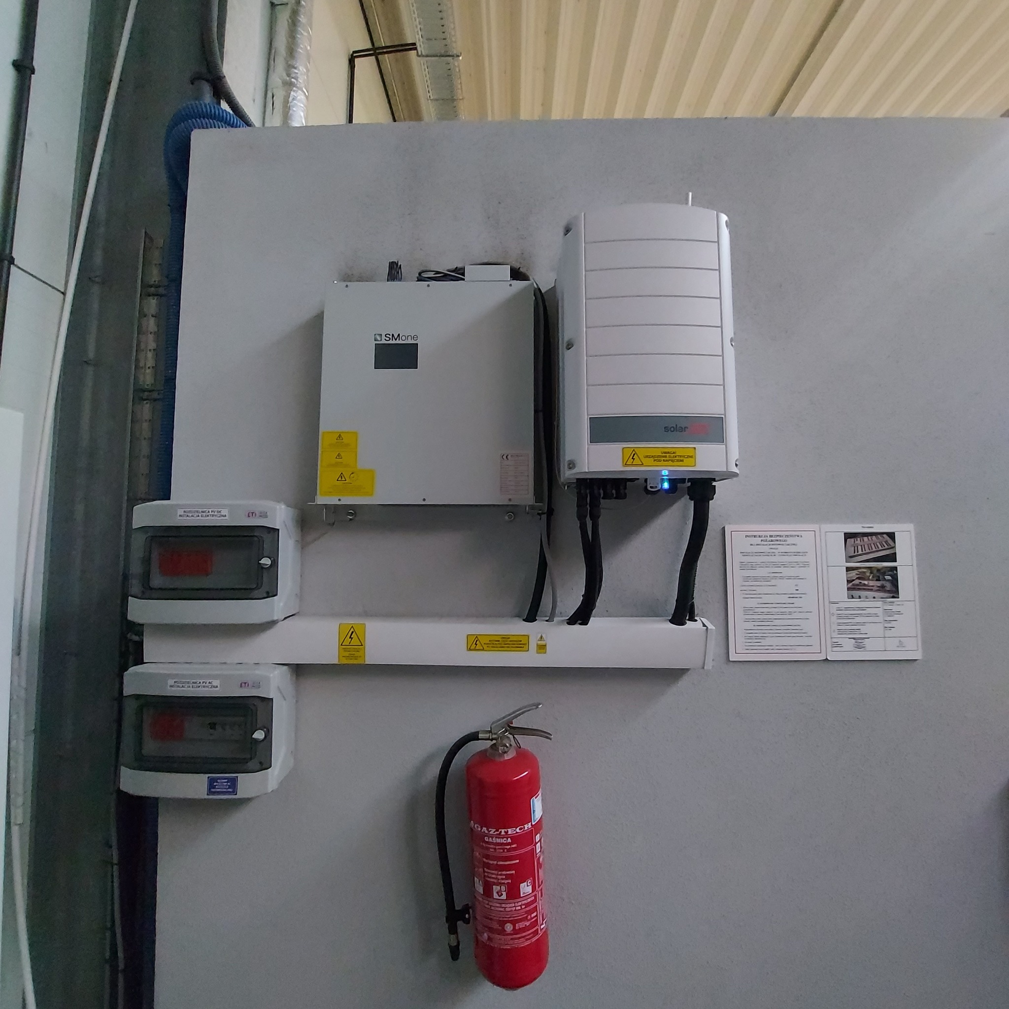Dwa inwertery, skrzynki bezpiecznikowe, gaśnica i dokumentacja techniczna zamontowane na szarej ścianie w pomieszczeniu technicznym. Widoczne żółte naklejki ostrzegawcze o napięciu elektrycznym.